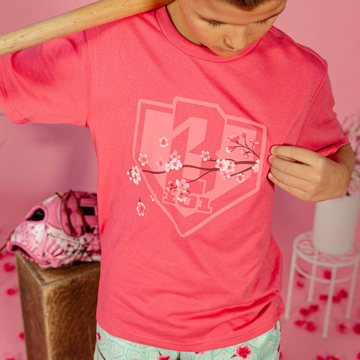Young boy holding a wooden bat over his shoulder showing the cherry blossom shield graphic on a pink BL101 tee against a pink background