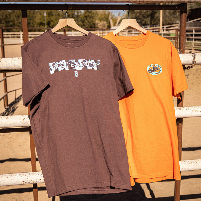 Two t-shirts, one brown with cow pattern and one orange, hanging on a wooden rack with a fence and trees in the background.