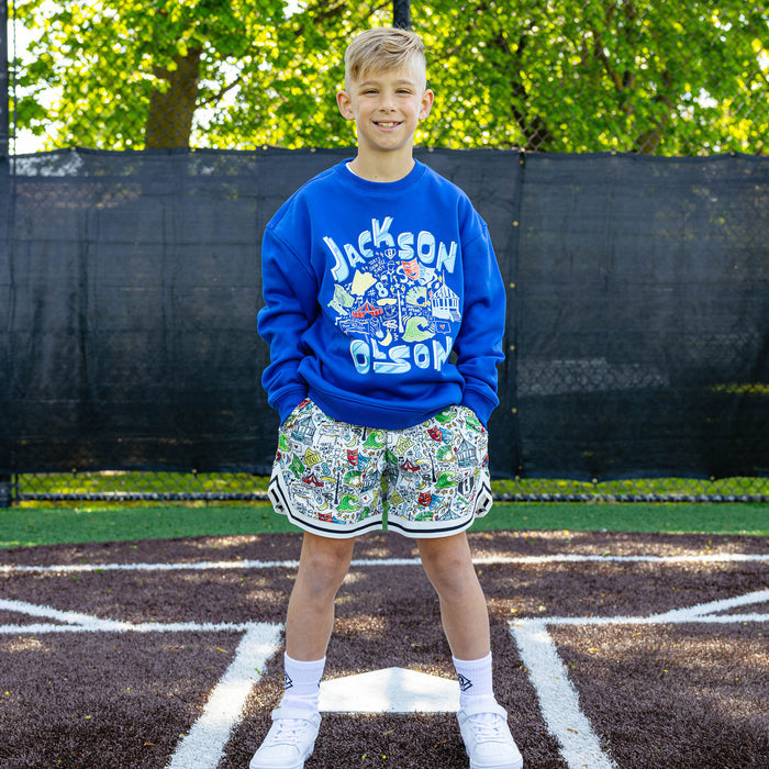 Smiling youth player at a baseball field wearing the royal blue Jackson Olson x BL101 Hometown Crewneck paired with the white Hometown Shorts