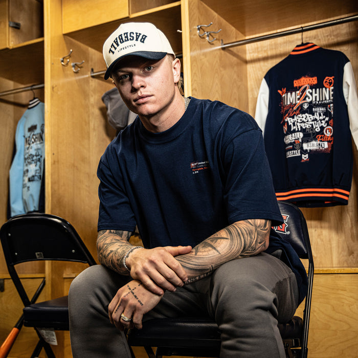 Person sitting in a locker room wearing a navy t-shirt and white cap, with tattoos visible.