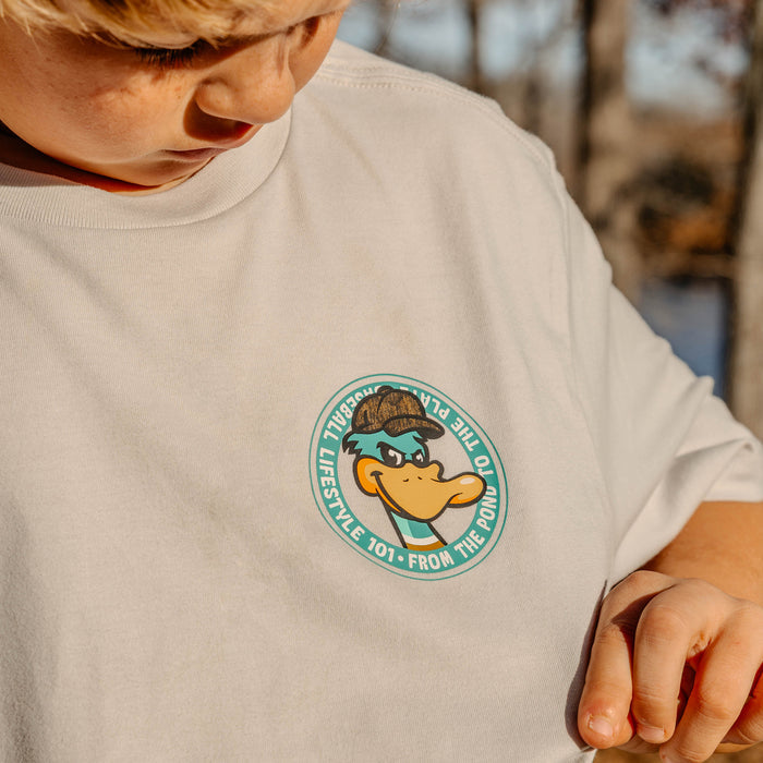Child wearing a light gray t-shirt with a duck character logo on a blurred natural background