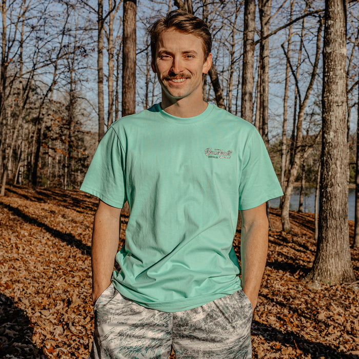 Man wearing a light green t-shirt with a Baseball Lifestyle 101 logo in a forest setting
