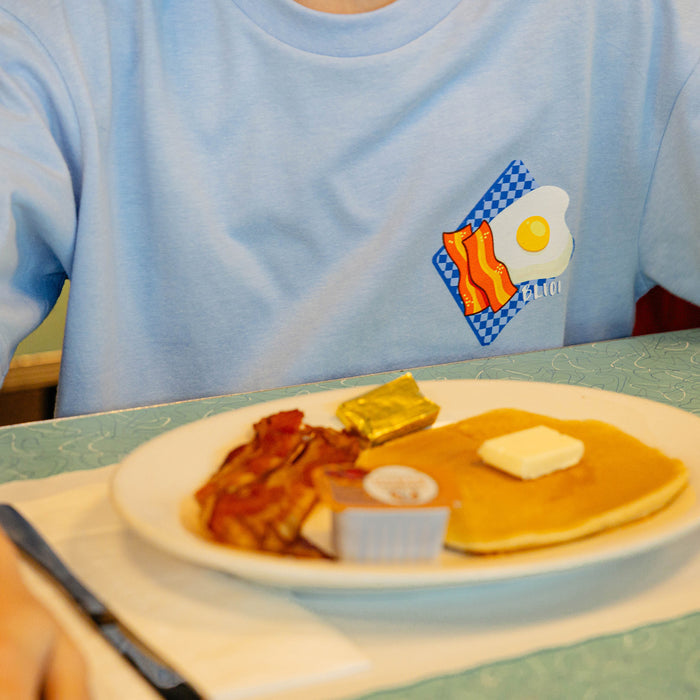 Person wearing a blue shirt with a bacon and egg graphic, sitting at a table with a plate of pancakes, bacon, and butter.