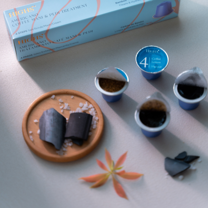 A box of Highs Americano Coffee Manicure and Pedicure Treatment with four capsules displayed in front of it, alongside a bowl of activated charcoal powder and a few dried leaves on a beige surface.