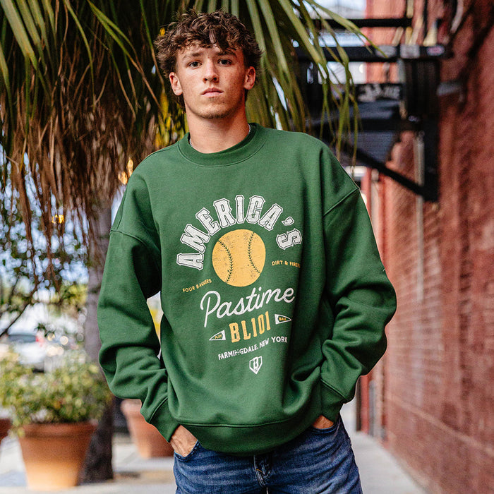Man with hands in pockets on a sidewalk wearing a forest green America's Pastime crewneck sweatshirt and jeans
