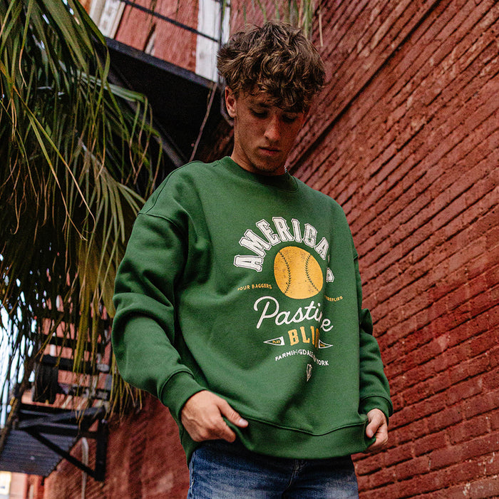 Man looking down against a brick wall wearing a forest green America's Pastime crewneck sweatshirt and jeans