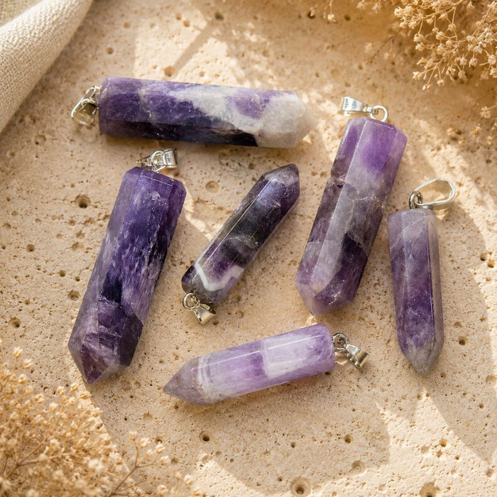 Five purple and white amethyst crystal point pendants with silver bail loops, arranged on a textured beige stone surface with dried floral props.