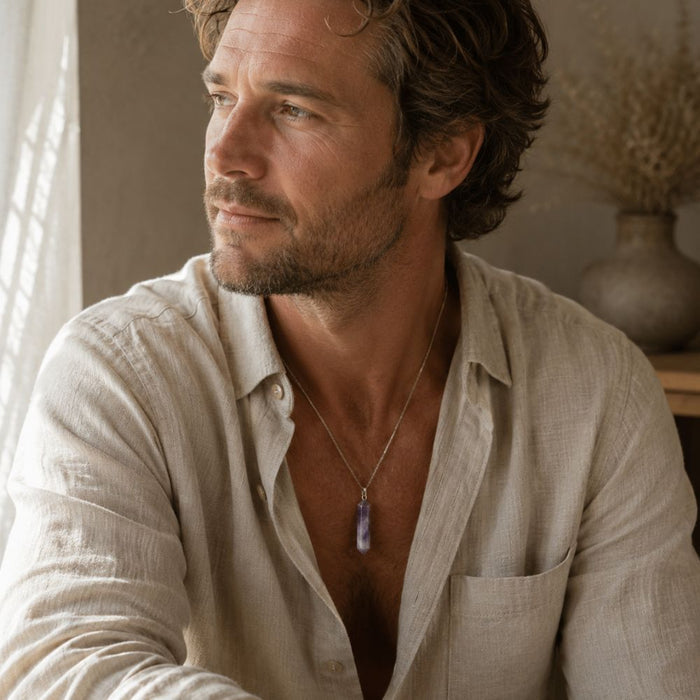 A man wearing a light beige linen button-up shirt and a silver pendant necklace with a purple stone, looking off-camera in a warm, sunlit room.