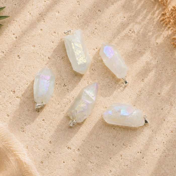 Aura clear quartz point pendants displayed on a warm natural stone surface, showing the unique shape, size, and iridescent finish of each crystal.