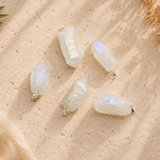Aura clear quartz point pendants displayed on a warm natural stone surface, showing the unique shape, size, and iridescent finish of each crystal.