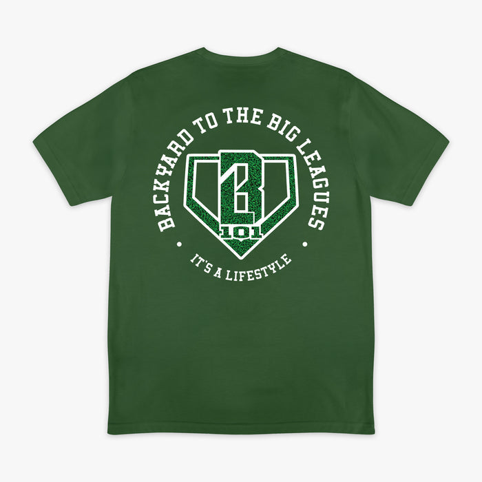 Image 1: Back flat lay of the BL101 Backyard to the Big Leagues forest green tee, featuring a circular "It's a Lifestyle" graphic with a grass-textured BL101 logo on a white background.