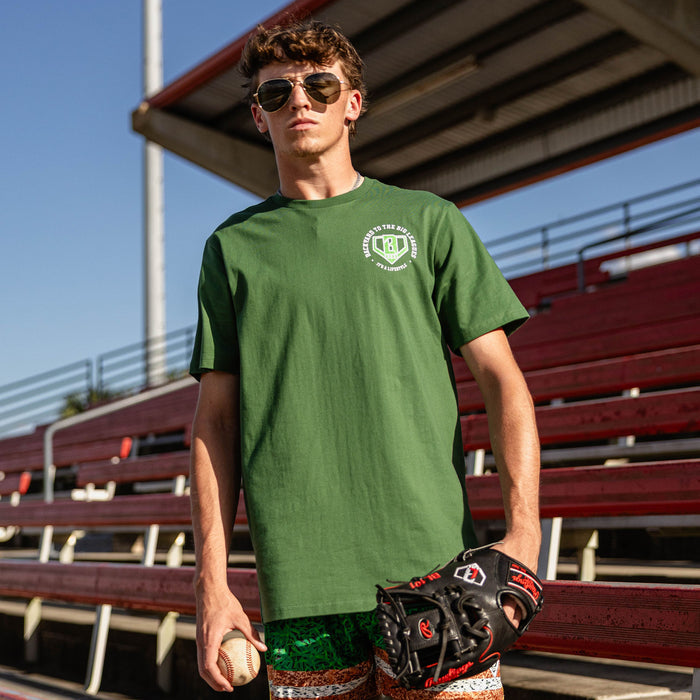 Man holding a baseball and glove at red stadium bleachers wearing aviator sunglasses and a forest green tee with a left chest BL101 logo graphic