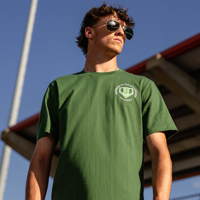 Man in aviator sunglasses looking up wearing a forest green tee with a left chest BL101 logo graphic against a blue sky
