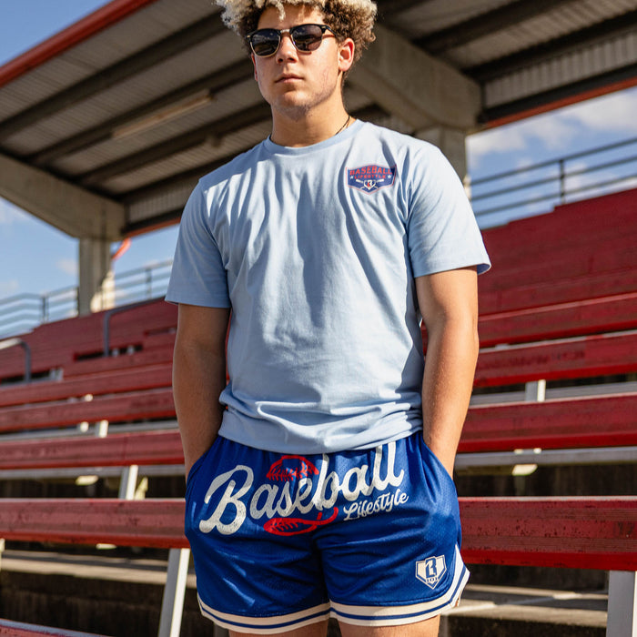 Man with curly hair in sunglasses standing at red stadium bleachers with hands in pockets wearing a light blue tee with a left chest BL101 logo graphic and royal blue Baseball Lifestyle mesh shorts