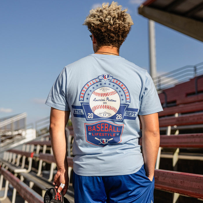Back view of man holding a glove at red stadium bleachers wearing a light blue tee with a Baseball Champions America's Pastime 101 back graphic and royal blue shorts