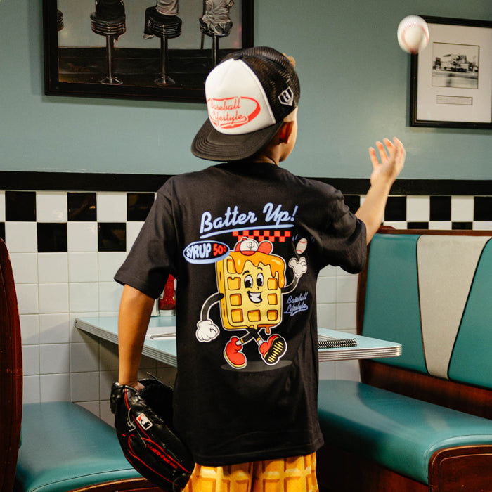 Child wearing a 'Batter Up!' t-shirt with a cartoon waffle design in a retro diner setting.