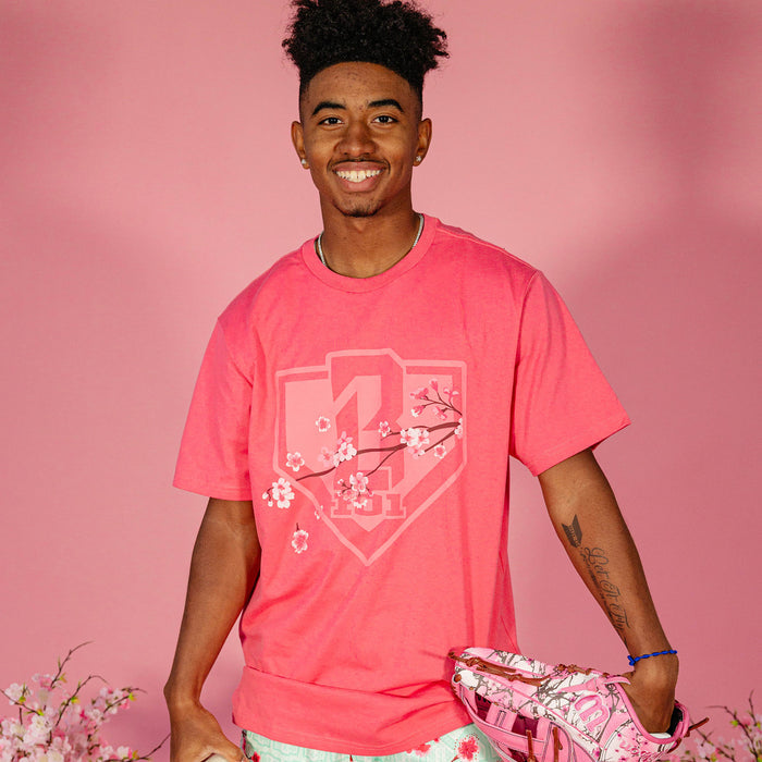 Male model wearing a pink BL101 cherry blossom shield tee and holding a pink floral baseball glove against a pink background, front view