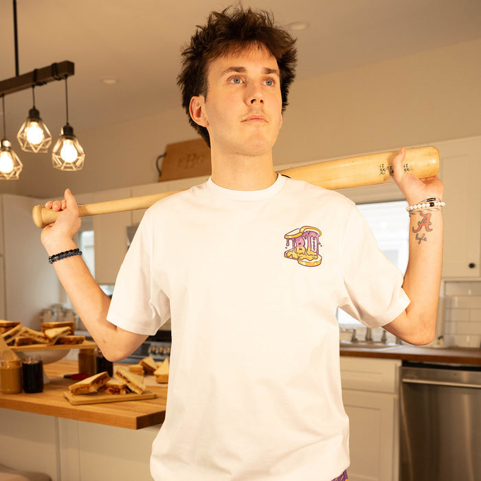 Male model holding a baseball bat in a kitchen wearing the BL101 Peanut Butter and Jelly white tee, showing the chest logo.