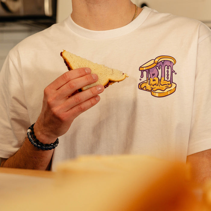 Close-up of the BL101 Peanut Butter and Jelly chest logo on the white tee, with a PB&J sandwich held in the foreground.