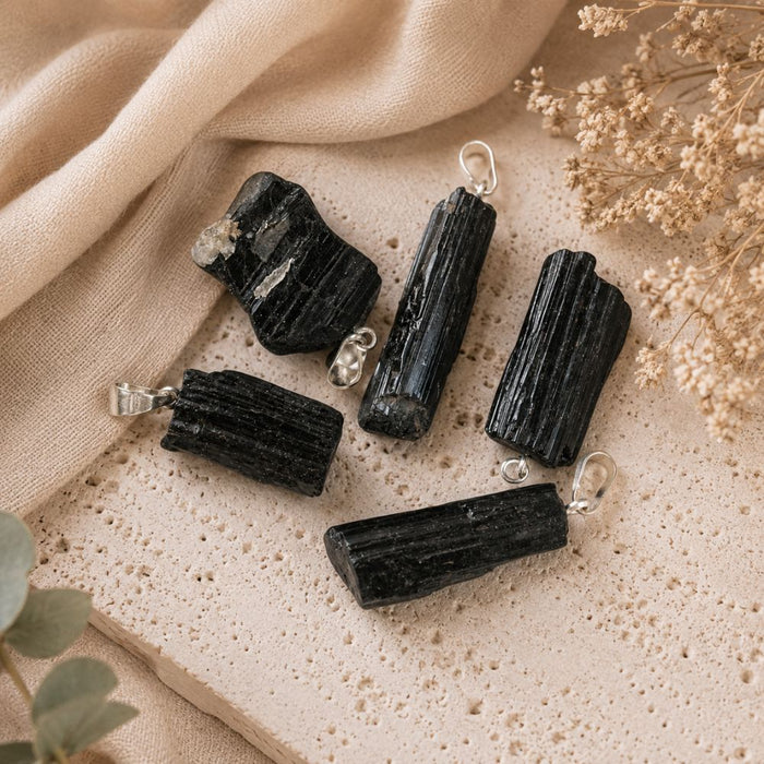Five black tourmaline crystal pendants with silver jump rings, arranged on a textured beige stone tile with draped beige fabric and dried flowers.
