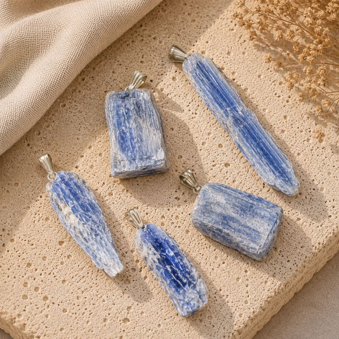 Five blue-and-white striped stone pendants with silver bail loops, arranged on a textured beige stone surface with a beige fabric and dried flowers in the background.
