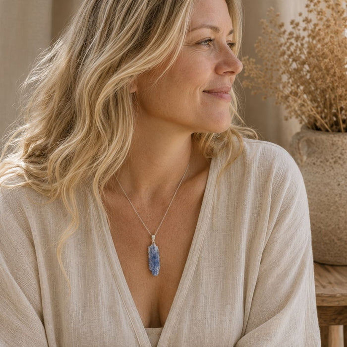 A woman with blonde hair wearing a silver pendant necklace with a blue-gray stone, dressed in a beige linen top, with dried flowers in the background.