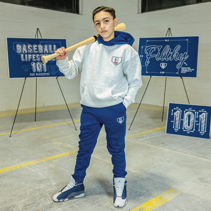 Person wearing a gray hoodie and blue pants holding a baseball bat in an indoor setting with 'Baseball Life 101' and 'Filthy' signs.