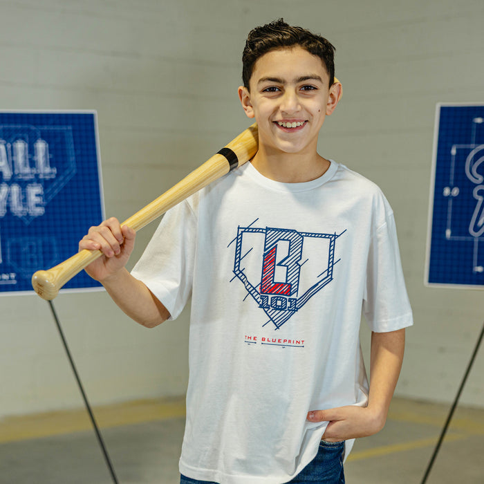 Young boy holding a baseball bat, wearing a white t-shirt with a BL101 logo, in an indoor setting.