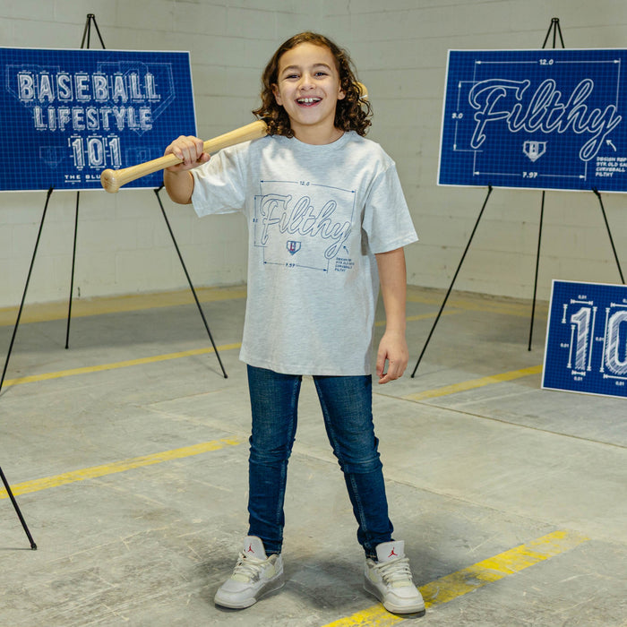 Child holding a baseball bat in front of 'Baseball Lifestyle 101' and 'Filthy' signs, wearing a gray t-shirt with blue 'Filthy' text and graphic design