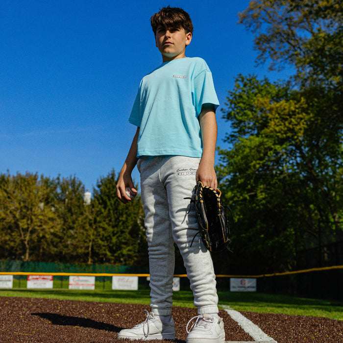Youth player standing on a baseball field holding a ball and glove, wearing the heather gray Jackson Olson x BL101 BSBL Era sweatpants with light blue tee
