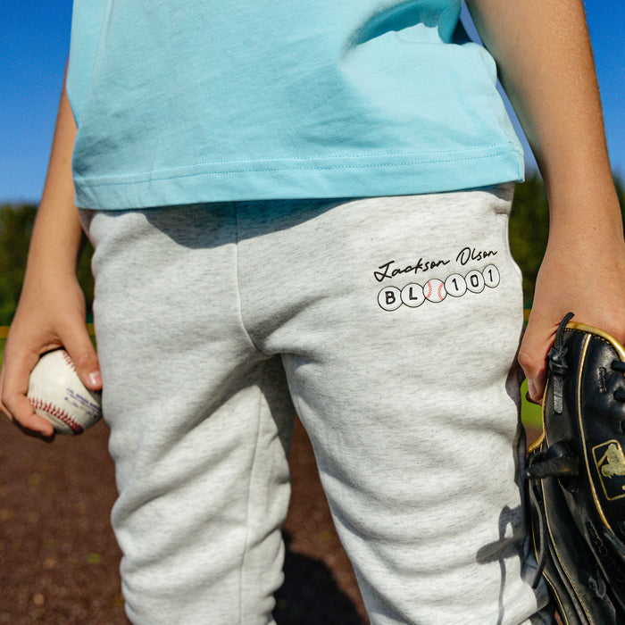 Close-up of the thigh graphic on the youth heather gray BSBL Era sweatpants, featuring the "Jackson Olson" script above the BL101 baseball bead logo