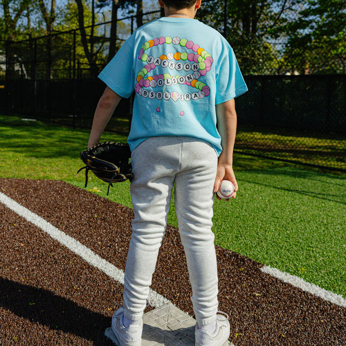 Back view of a youth player on a baseball field wearing the heather gray BSBL Era sweatpants paired with the light blue BSBL Era tee showing the friendship bracelet back graphic