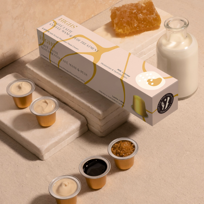A boxed manicure and pedicure treatment product with capsules for different steps of the process laid out next to it, with a background of milk and honey containers and a stack of coffee-colored ceramic cups.
