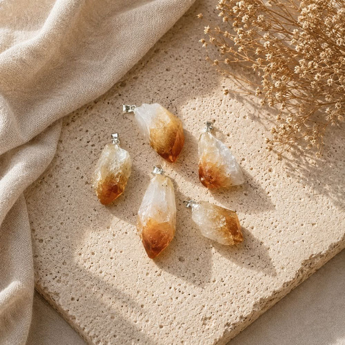 Four natural citrine crystal point pendants with silver bail loops, arranged on a textured beige stone slab with draped beige fabric and dried flowers.