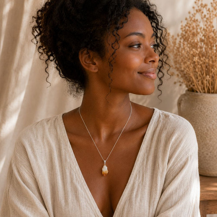 A woman with curly dark hair wearing a silver pendant necklace with a natural cowrie shell pendant, posing in a light beige linen top against a warm, softly lit background with dried flowers.