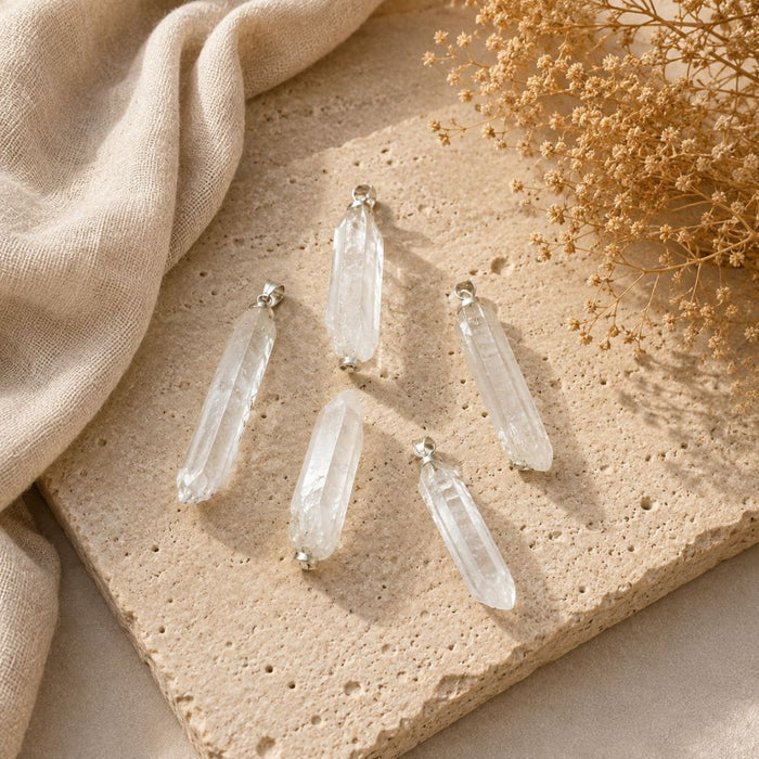 Five clear crystal point pendants with silver-toned caps arranged on a beige stone slab with a beige fabric and dried flowers in the background.