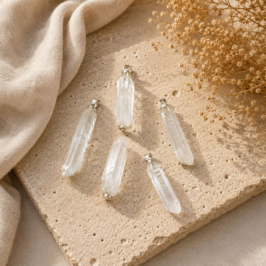 Five clear crystal point pendants with silver-toned caps arranged on a beige stone slab with a beige fabric and dried flowers in the background.
