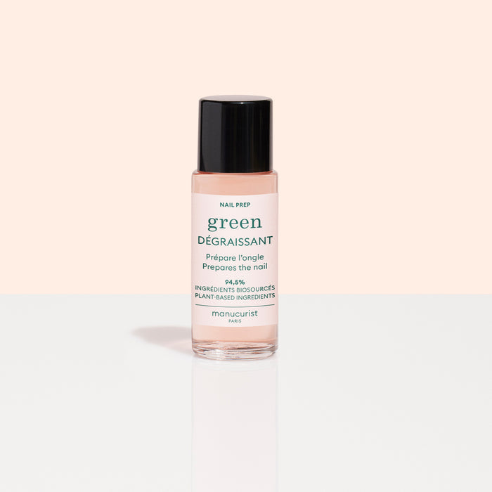 A small bottle of Manucurist Degreaser, a plant-based nail prep with clear pink liquid and black cap, sits on a reflective white surface. Its white label with green and pink text stands out against a pale peach background.
