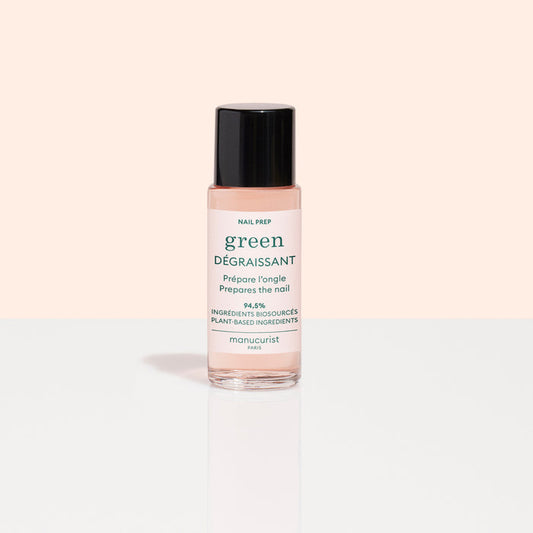 A small bottle of Manucurist Degreaser, a plant-based nail prep with clear pink liquid and black cap, sits on a reflective white surface. Its white label with green and pink text stands out against a pale peach background.
