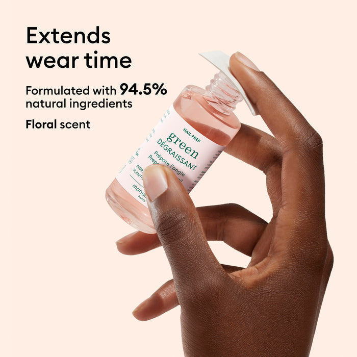 A hand holds a pink Manucurist Degreaser bottle labeled green against a beige background. Text: Extends nail polish adhesion. Made with 94.5% natural ingredients, this plant-based nail product features a floral scent.