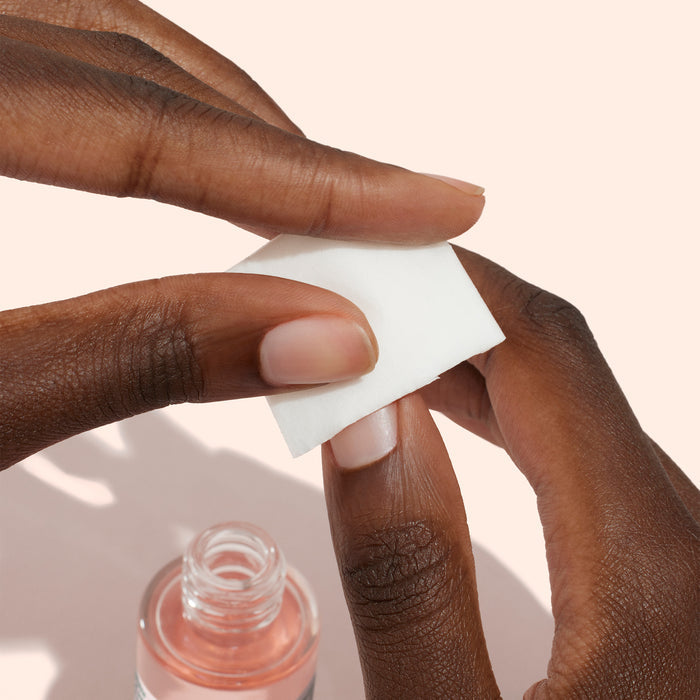 A person uses a white cotton pad to clean their fingernail with Manucurist Degreaser, a plant-based nail prep product that enhances polish adhesion, shown next to the bottle against a light background.