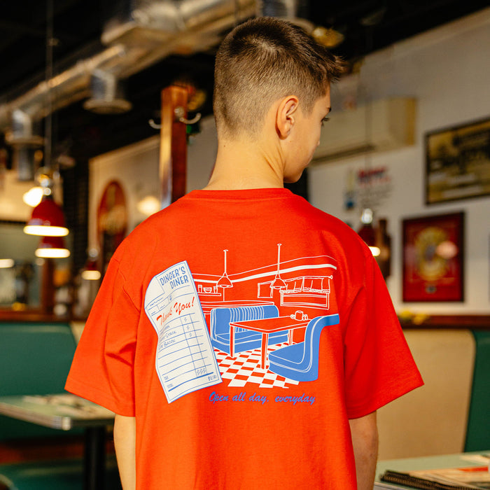 Person wearing a red t-shirt with a graphic design on the back featuring a restaurant scene and checkered floor.