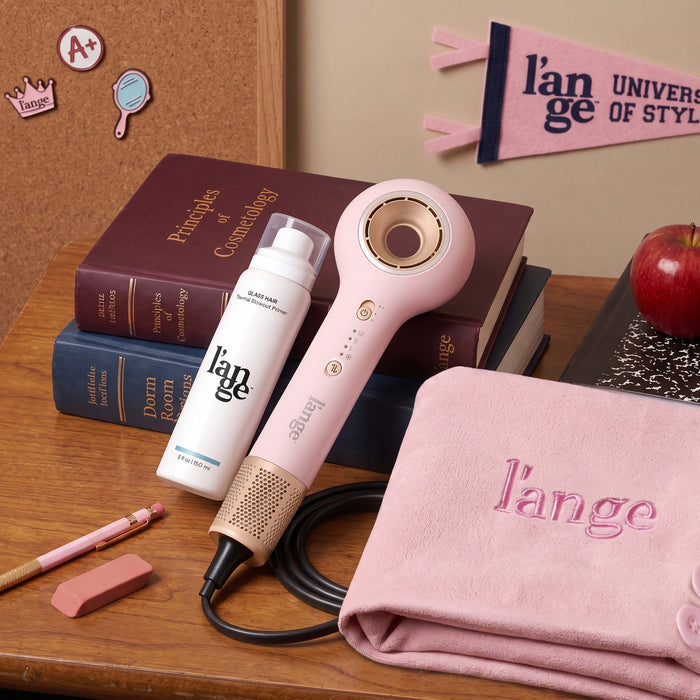 Axia hair dryer set on a wooden table, alongside a light pink hair towel and Glass Hair Primer, surrounded by books and school supplies.