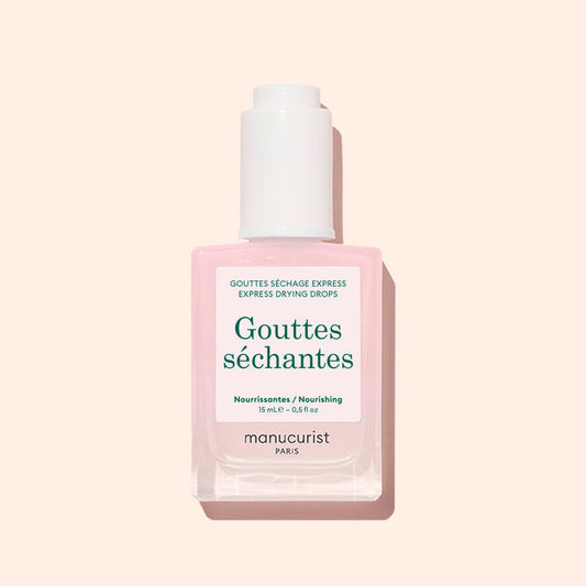 A square bottle of Manucurist Drying Drops with a white cap, featuring light pink liquid, is shown against a pale pink background. This nail product speeds up drying time and nourishes cuticles.