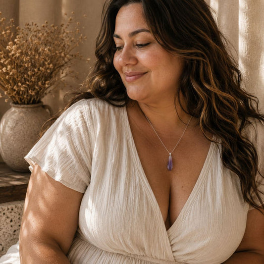 A woman with long wavy brown hair wearing a cream-colored crinkled wrap dress and a silver pendant necklace with a purple stone, set in a warm, sunlit room with dried flowers in the background.