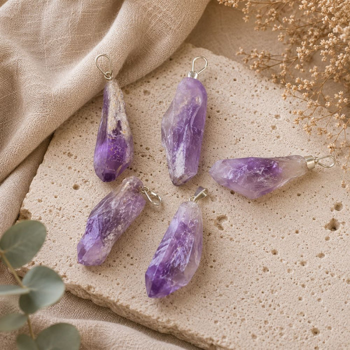 Four raw amethyst crystal pendants with silver wire loops, arranged on a textured beige stone slab with beige fabric and dried flowers.