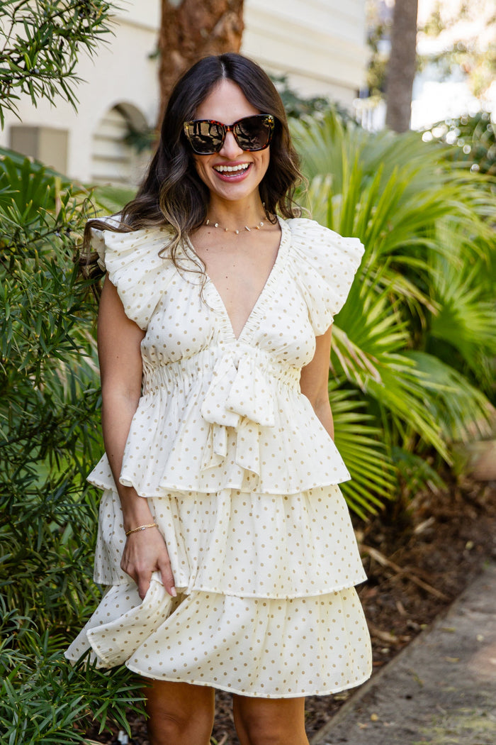 Woman in a layered white dress standing outdoors with greenery and a building in the background