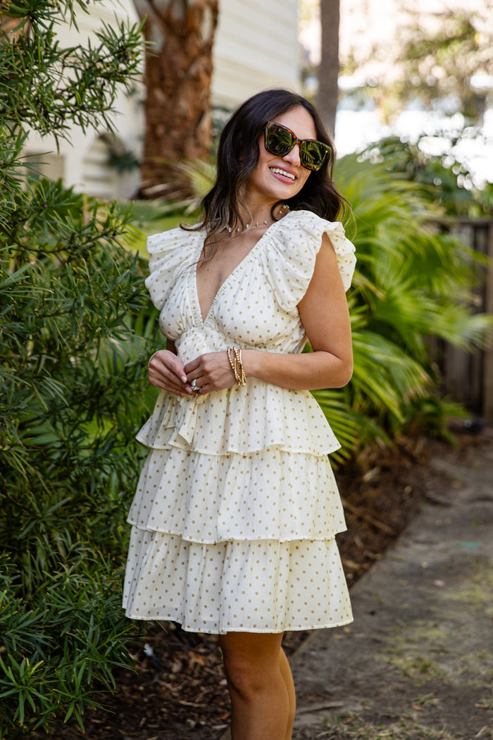 Woman in a white polka dot dress standing outdoors with greenery in the background