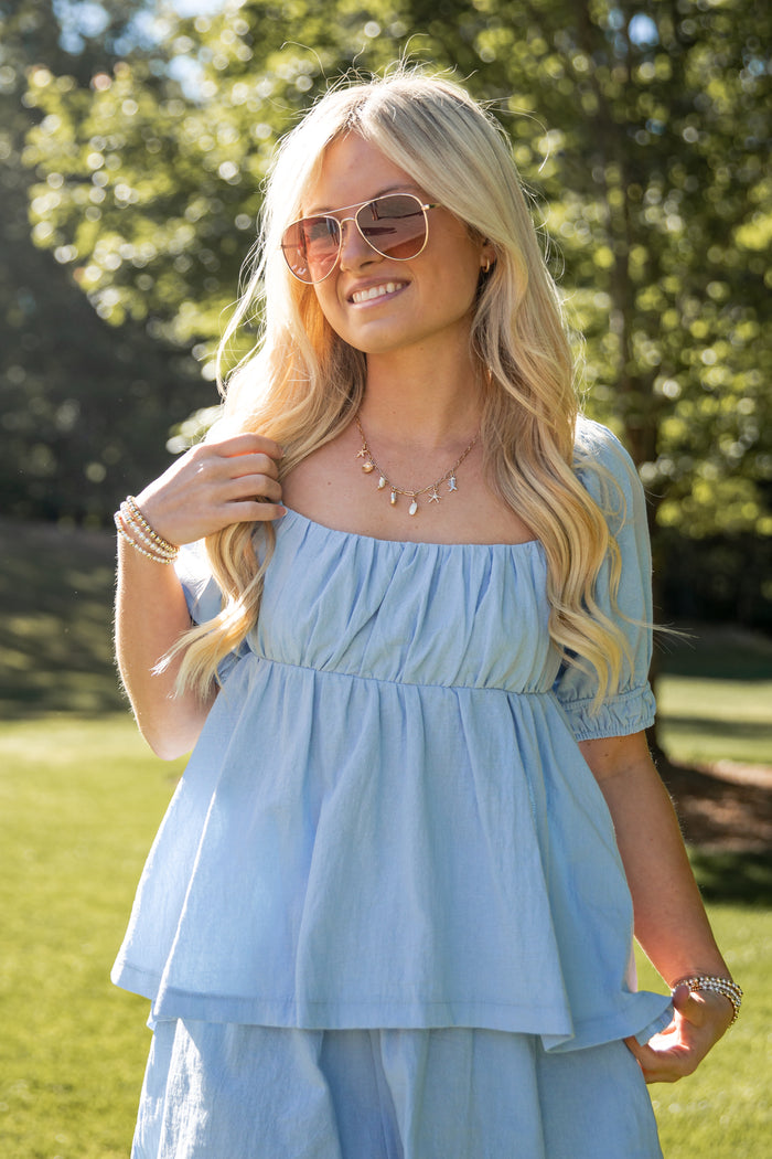 Woman wearing a light blue dress and sunglasses outdoors with trees in the background
