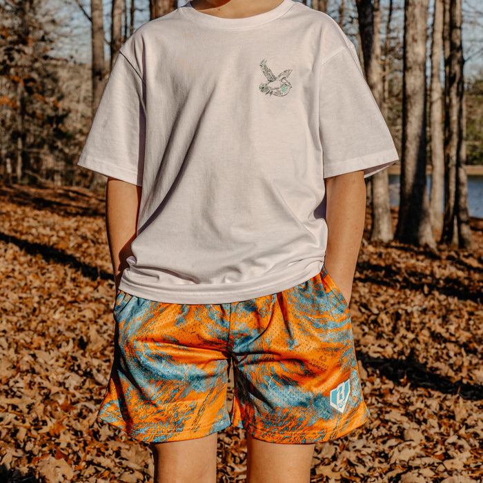 Person wearing a white t-shirt with a small bird design and orange and blue shorts in an outdoor setting with trees and fallen leaves.